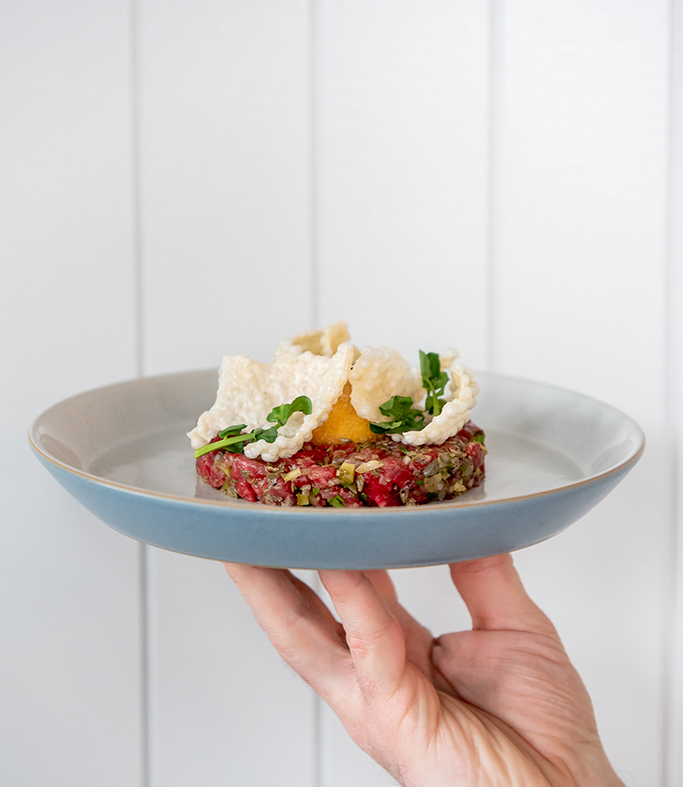 Hand holding Wagyu Tartare on a blue plate against a white backdrop